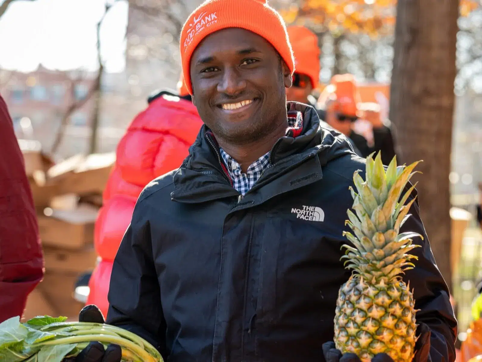 Smiling man in black jacket and orange Food Bank beanie holding a pineapple and leafy greens at an outdoor food distribution event.